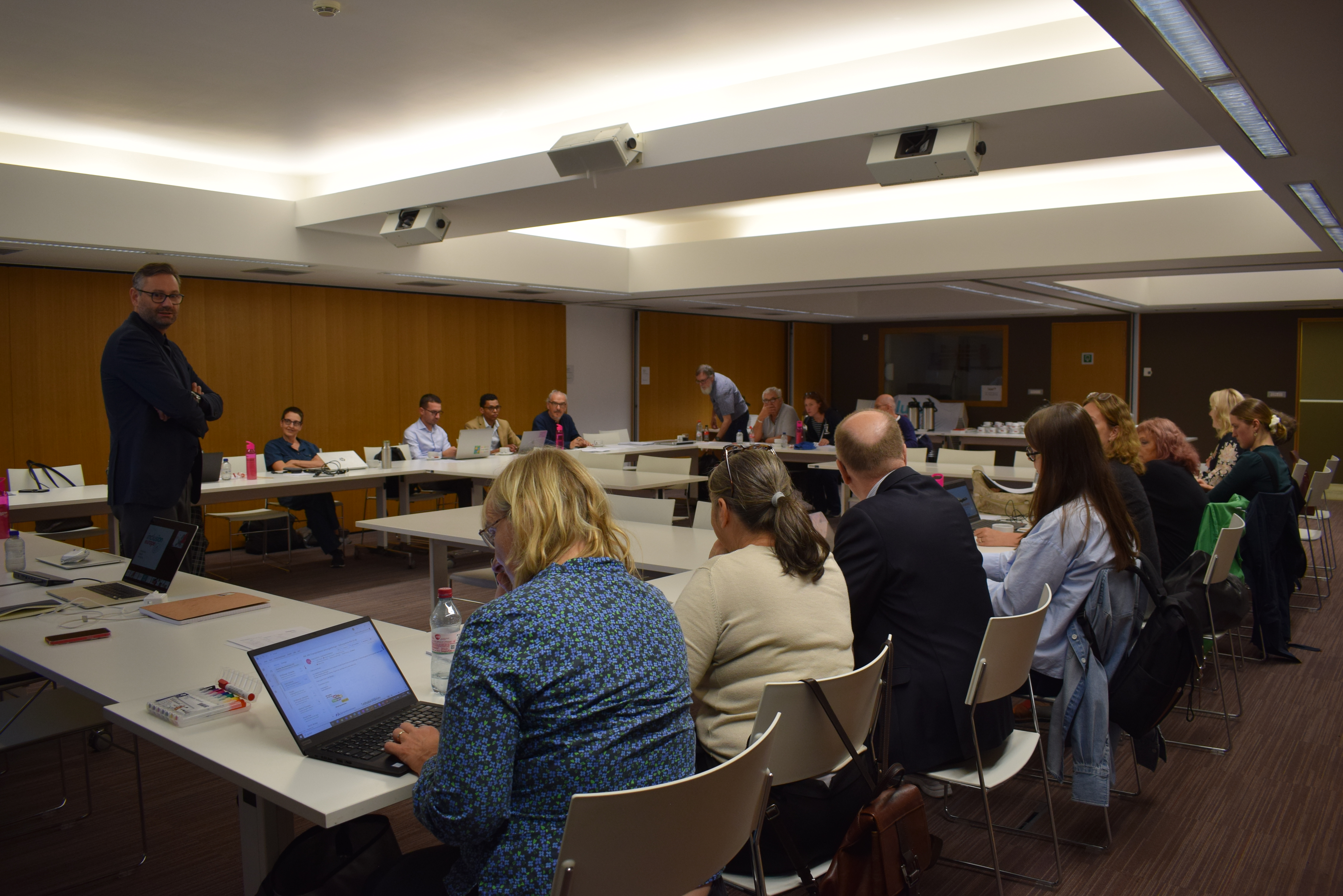 Participants sitting in the conference room around a big table and a moderator standing and speaking
